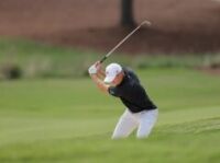 The Best Golf Books To Improve Your Game Golfer in a white cap and black shirt mid-swing on a green golf course fairway.