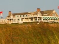 Father’s Day Golf Gifts to Save Dad a Trip to the Pro Shop Coastal golf clubouse on a hill, with white porch, green-and-white striped awning, flags, and people on the grass in the foreground.