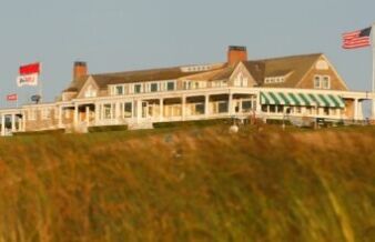 Father’s Day Golf Gifts to Save Dad a Trip to the Pro Shop Coastal golf clubouse on a hill, with white porch, green-and-white striped awning, flags, and people on the grass in the foreground.