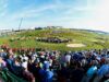 Amateur Golfers Set to Compete in the 2026 US Open Crowd of spectators fills a hillside golf course during a sunny tournament, with grandstands on the left and players near the green beneath a blue sky.