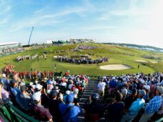 Amateur Golfers Set to Compete in the 2026 US Open Crowd of spectators fills a hillside golf course during a sunny tournament, with grandstands on the left and players near the green beneath a blue sky.