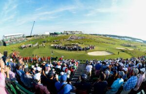 Amateur Golfers Set to Compete in the 2026 US Open Crowd of spectators fills a hillside golf course during a sunny tournament, with grandstands on the left and players near the green beneath a blue sky.