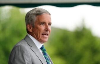 Jay Monahan Unloads on Team USA’s Curtis Cup Challenges Older man in a light gray suit, seen in profile, speaking outdoors with a blurred green background.