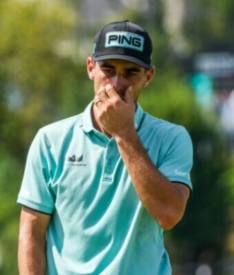 Joaquin Niemann Receives Two-Stroke Penalty for ‘Serious Misconduct’ at US Open Male golfer in a turquoise polo shirt and black Ping cap touches his face while standing outdoors on a sunny golf course behind him blurred trees and spectators