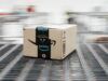 30+ Of The Best Amazon Prime Day Golf Deals I Have Spotted In 2026 Brown Amazon cardboard box on a moving conveyor belt in a warehouse, blurred background conveys motion
