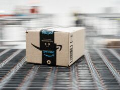 30+ Of The Best Amazon Prime Day Golf Deals I Have Spotted In 2026 Brown Amazon cardboard box on a moving conveyor belt in a warehouse, blurred background conveys motion
