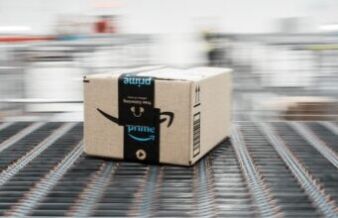 30+ Of The Best Amazon Prime Day Golf Deals I Have Spotted In 2026 Brown Amazon cardboard box on a moving conveyor belt in a warehouse, blurred background conveys motion