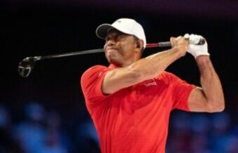 Tiger Woods Introduces Major PGA Tour Reforms in Rare Appearance Male golfer in a red polo shirt and white cap mid-swing, gripping a golf club against a dark background.