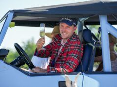 Russell Henley Drops 15-Club Bag to Win at Charles Schwab Man in a red plaid shirt and Titleist cap raises a champagne flute while sitting in a light-blue convertible, smiling on a sunny day.