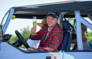 Russell Henley Drops 15-Club Bag to Win at Charles Schwab Man in a red plaid shirt and Titleist cap raises a champagne flute while sitting in a light-blue convertible, smiling on a sunny day.