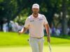 Clark Triumphs with Resilience at US Open Golfer walking on a sunny green course, wearing a pale pink polo and a white cap with a T-Mobile logo, holding a golf club.