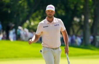 Clark Triumphs with Resilience at US Open Golfer walking on a sunny green course, wearing a pale pink polo and a white cap with a T-Mobile logo, holding a golf club.
