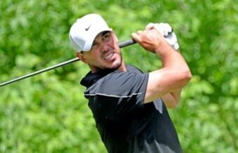 What Brooks Koepka’s Withdrawal Actually Means for the US Open Male golfer in a black shirt executing a backswing with a golf club, white Nike cap, light glove, on a lush green backdrop.