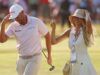 Clark Sets 36-Hole U.S. Open Shinnecock Record Man in a white polo shirt adjusts his cap while walking beside a smiling woman in a sunhat at a sunny golf event.