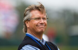 Scotty Cameron GOLO 6 Makes Traditional Putters Obsolete Smiling middle-aged man with gray hair and glasses outdoors, wearing a light blue shirt and dark vest with a blue lanyard.