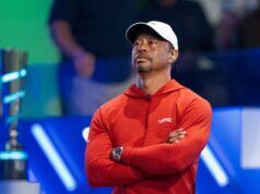 Tiger Woods Sets Unbreakable 142-Cut Streak Record Man in a red hoodie with a white cap, arms crossed, listening on a stage with blue background lights.