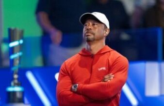 Tiger Woods Sets Unbreakable 142-Cut Streak Record Man in a red hoodie with a white cap, arms crossed, listening on a stage with blue background lights.