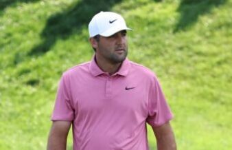 Scheffler Stuns Field as Scottish Open Showdown Begins Man wearing a pink polo shirt and white Nike cap standing in a grassy outdoor area with greenery in the background.