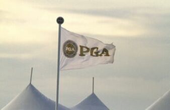 PGA Tour Legend And Eight-Time Winner Dead at 85 White flag bearing a circular PGA logo waves atop a pole beside white event tents under a pale sky.