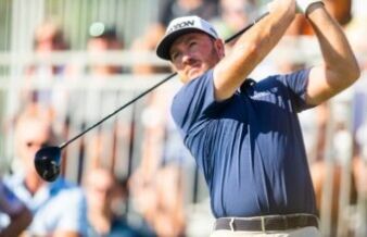 LIV Golf Star Admits Why He Joined Controversial League Male golfer in a navy shirt swings a club during a sunny tournament, crowd blurred in the background at the course.
