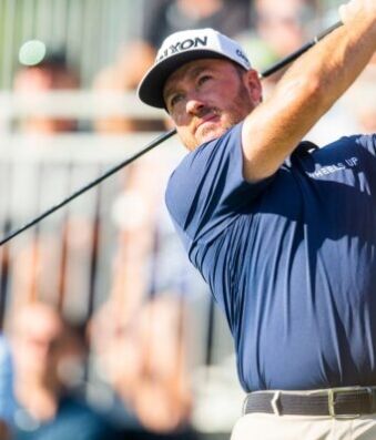 LIV Golf Star Admits Why He Joined Controversial League Male golfer in a navy shirt swings a club during a sunny tournament, crowd blurred in the background at the course.