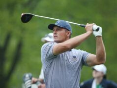 Bryson DeChambeau Lays Out 2 Demands for PGA Tour Return Golfer in a blue cap and striped shirt mid-swing on a golf course, glove on his left hand.