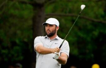 Scottie Scheffler Gives Sassy Response To LIV Golf Question Male golfer in a white cap and polo swinging a golf club on a grassy course, focused on the shot.