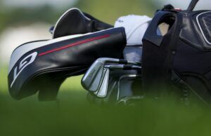 A 10-Handicap and a +4-Handicap Got Fitted—Then This Happened Close-up of a golf bag with a Titleist iron resting beside it on a blurred green background, highlighting black and white bag details with a red stripe.