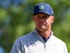 Bryson DeChambeau Just Admitted He Doesn’t Know What To Do Head-and-shoulders portrait of a man outdoors wearing a blue cap and a light gray zip-up sweater; patch on the cap shows a white skull and crossed bones, blurred green background.