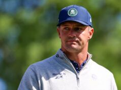 Bryson DeChambeau Just Admitted He Doesn’t Know What To Do Head-and-shoulders portrait of a man outdoors wearing a blue cap and a light gray zip-up sweater; patch on the cap shows a white skull and crossed bones, blurred green background.
