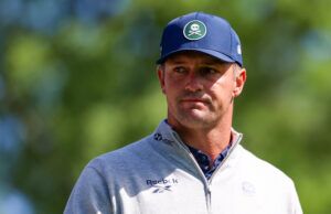 Bryson DeChambeau Just Admitted He Doesn’t Know What To Do Head-and-shoulders portrait of a man outdoors wearing a blue cap and a light gray zip-up sweater; patch on the cap shows a white skull and crossed bones, blurred green background.