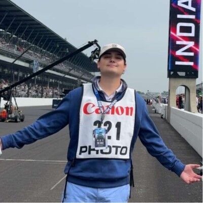Person wearing a Canon photo bib with number 21 and a cap, arms outstretched on a race track with grandstands and an INDY sign behind.